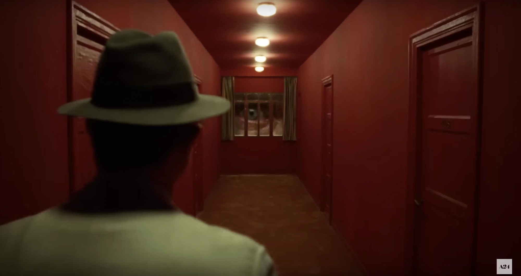 A man walks through a red hallway. At the end of the hallway is window that is fill by a giant eye.