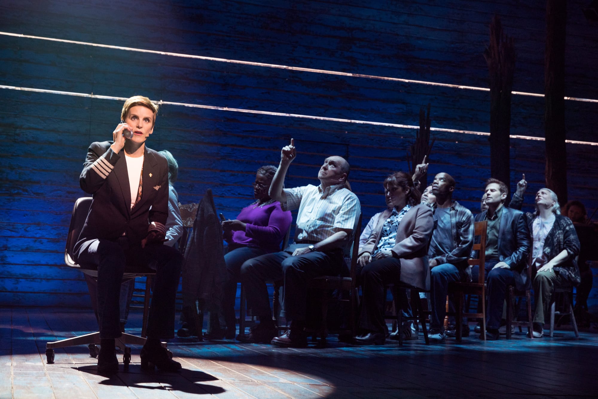 Onstage, in the spot light,a concerned-looking flight attendant speaking into an intercom receiver. To her right, people in two rows of chairs seated like airline passengers, some of them pressing up at the buttons on the imagined ceiling above them.