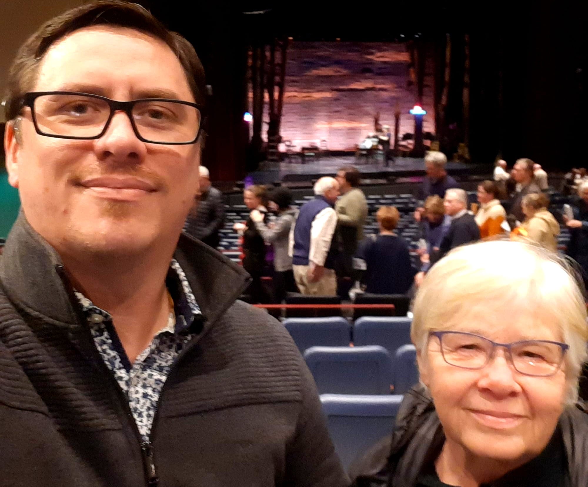 To the left, the author, allowing a smile. To the right and a foot lower, his mother. Behind them a departing theater audience, with the stage lit further back..