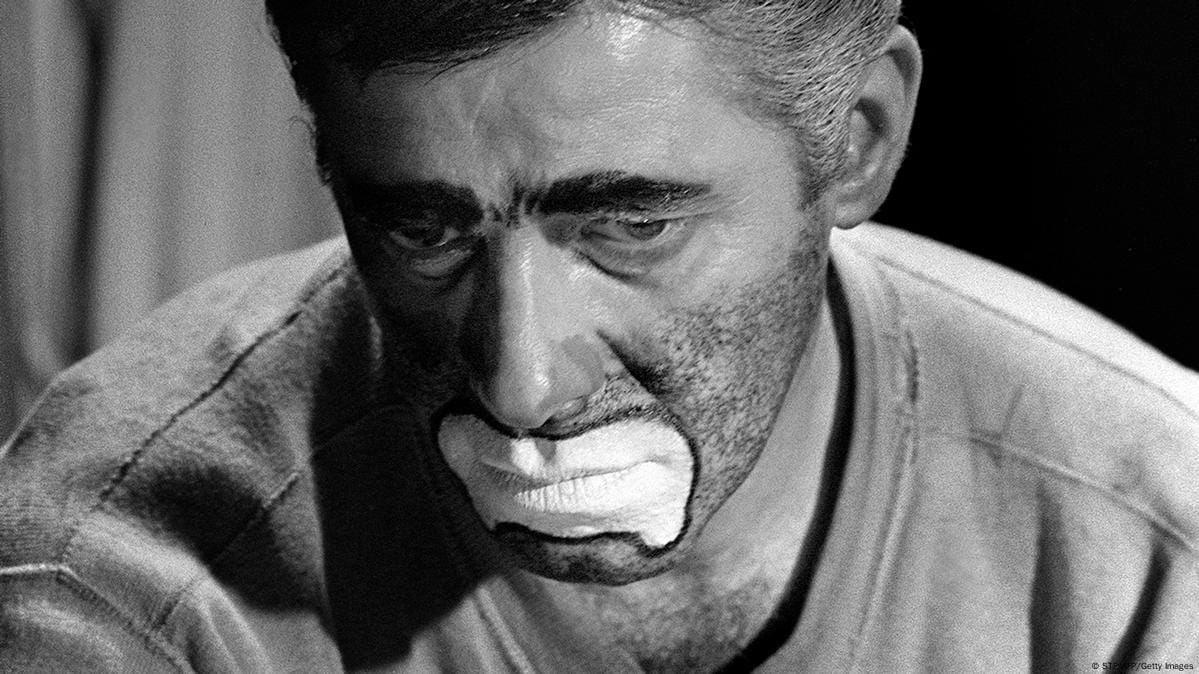 In black and white, a close-up of a middle-aged man with frowning white clown face paint.