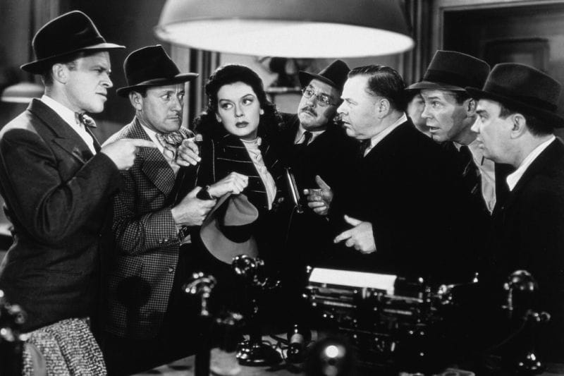 In black-and-white, six men surround a woman, waiting on her response. She does not look interested in giving them one.