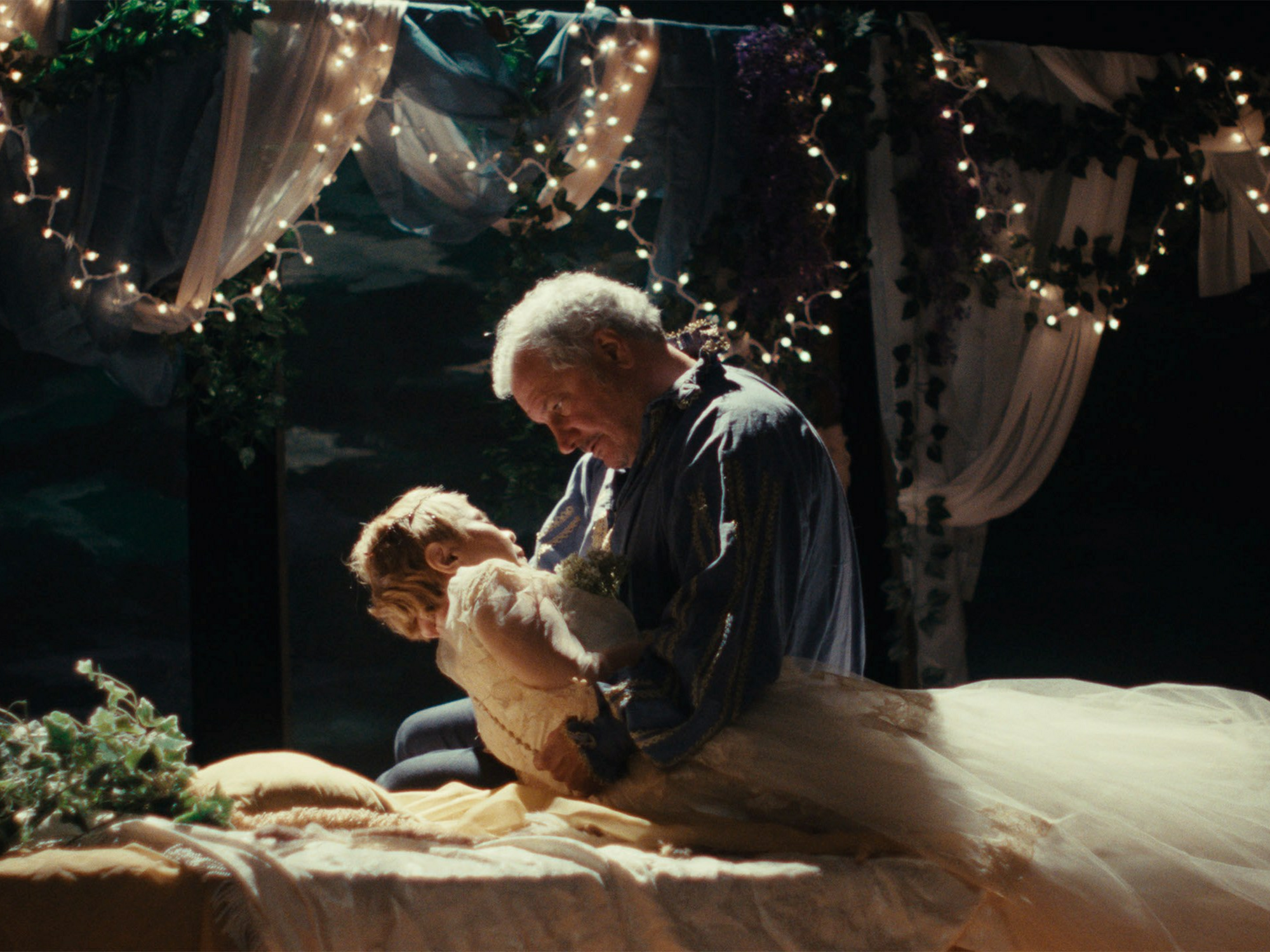 A middle-aged man in Renaissance garb sits at a bed and holds a woman in a wedding dress.The bedposts are decorated in strings of lights and semi-translucent sheets.