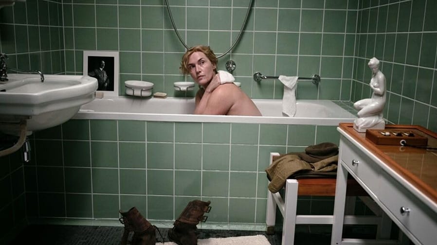 A woman in a bathtub, her arms crossed to cover her breasts, looking over her shoulder. Her pose is modeled after a Venus-like statuette to the right. To the left, a framed photo of Adolf Hitler. Outside the tub dirty boots stand on a towel.