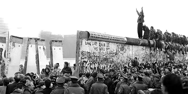 Sections of the Berline wall are removed, while several young people sit atop, their legs straddling either side. One of them at the front stands, his arms waving high in jubilation.