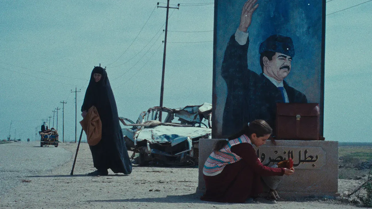 A young girl kneels and feeds her pet rooster in front of a standalone mural of Saddam Hussein. Behind her, in front of a burned-out car on the side of the road, an elderly woman in a chador supports herself with a cane.