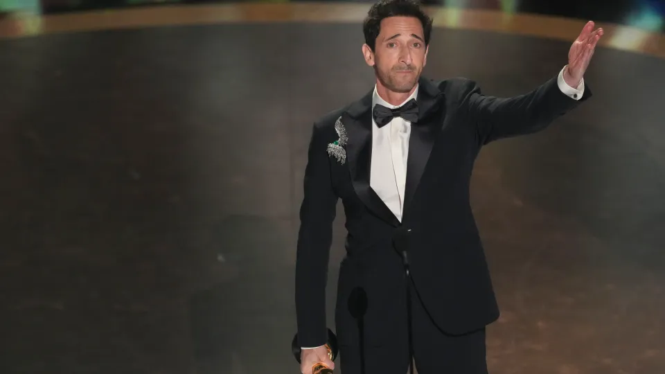 Adrian Brody stands pensively on the Oscars stage, his left arm extended, his right arm clutching his prize statuette..